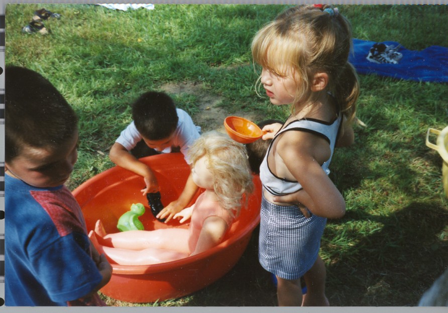 Drie kinderen spelen buiten met een grote pop in een oranje emmer vol water op het gras.
