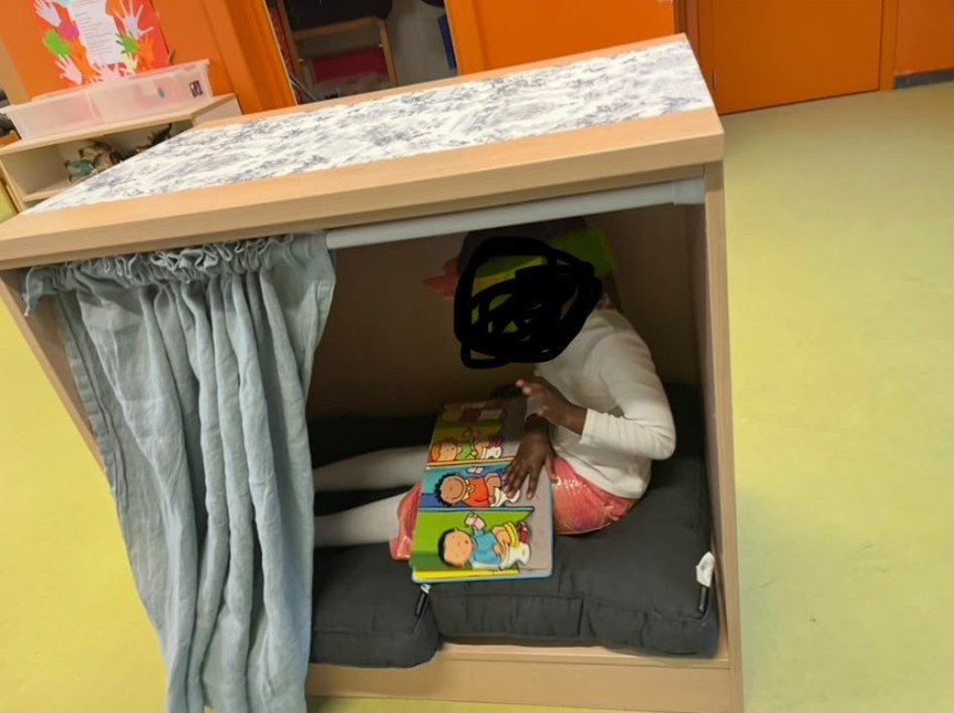 Kind zit in een leeshuisje op kussen en leest een boek met kleurrijke illustraties.