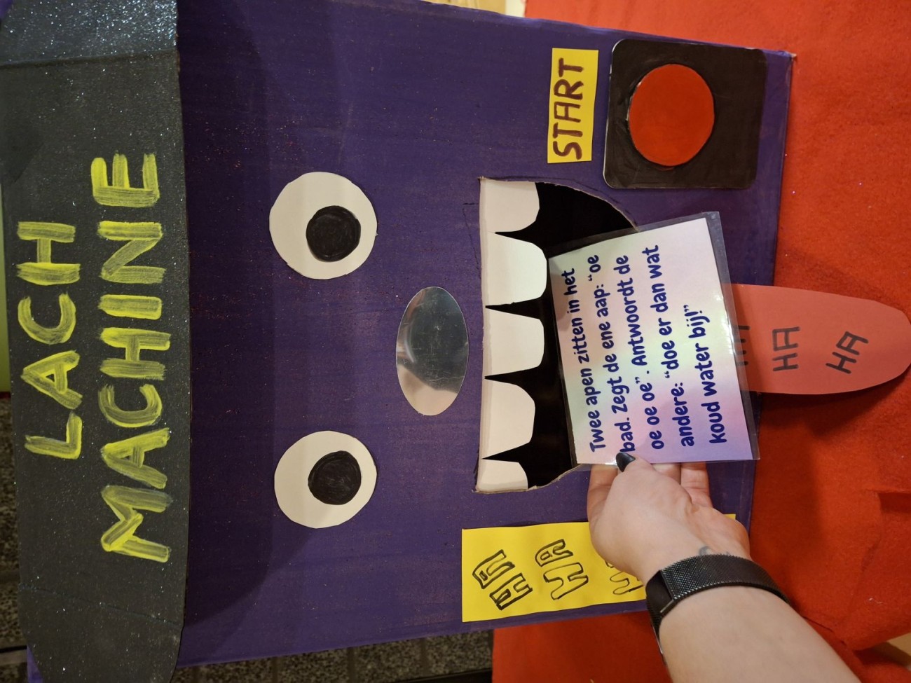 Hand plakt tekst in mond van morse lachmachine met grote ogen, tanden en tong, op rode achtergrond.