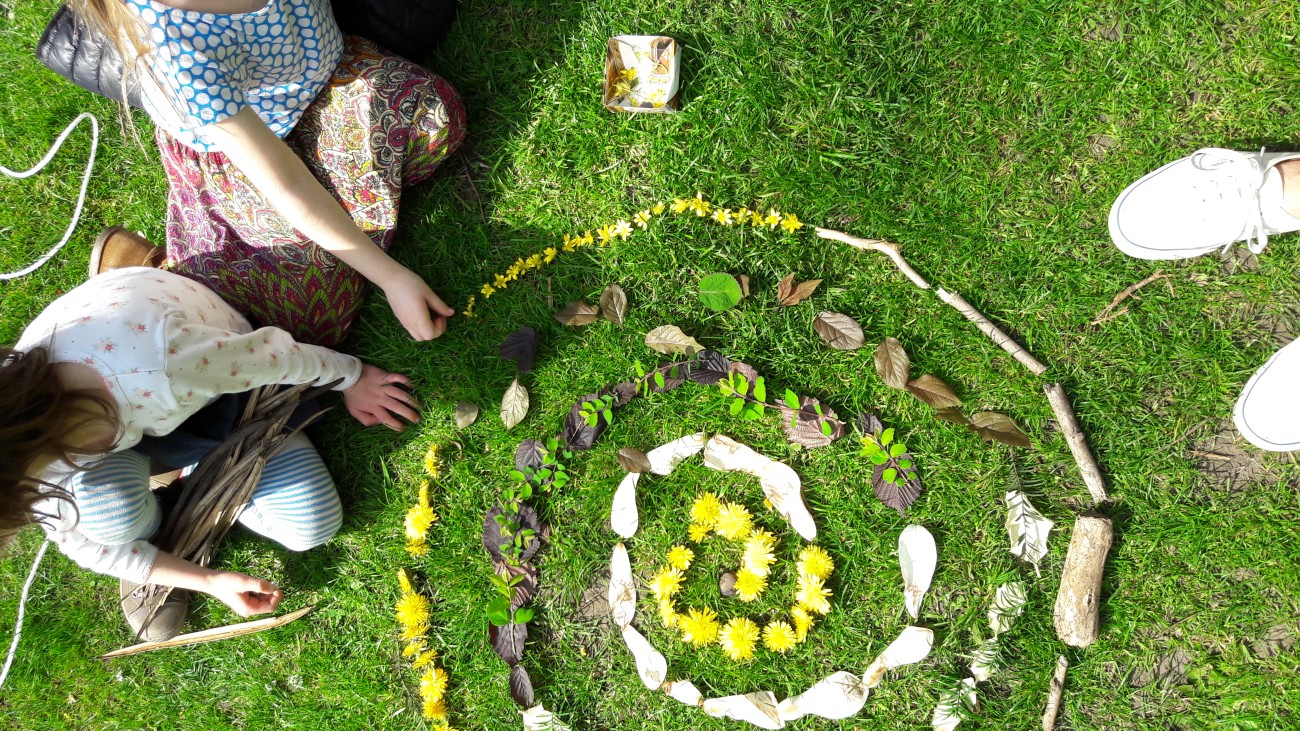 Twee kinderen maken een spiraalvormige tekening op gras met bloemen, bladeren en takken.