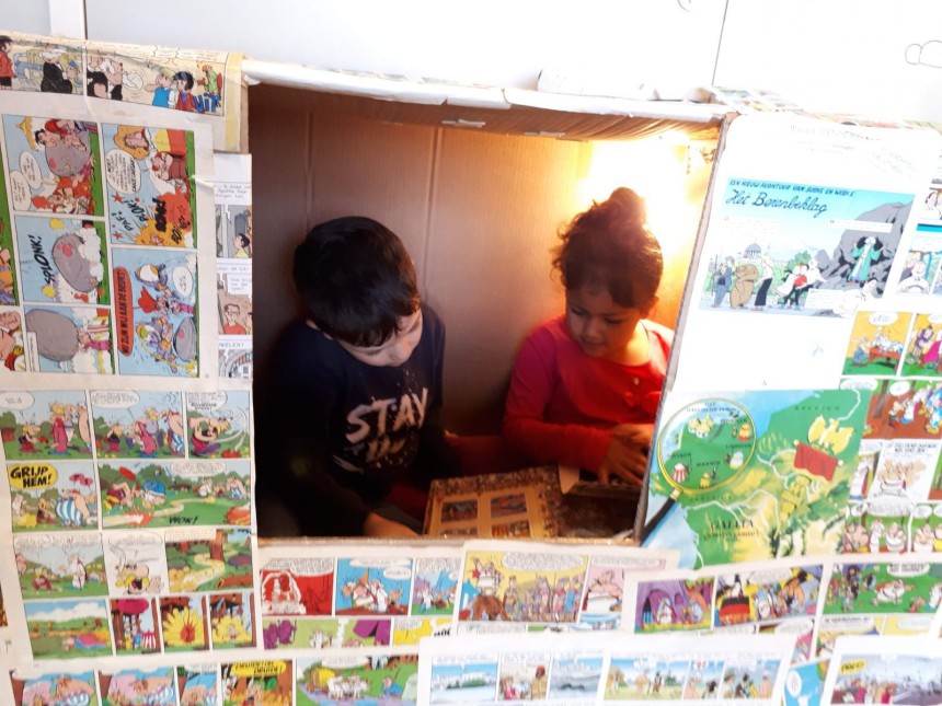 Twee kinderen zitten samen in een kartonnen doos bekleed met kleurrijke strips.