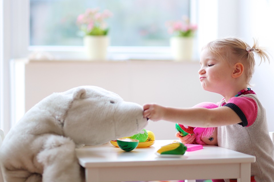 Een meisje speelt met een grote witte knuffelbeer aan een kleine tafel in een lichte kamer.