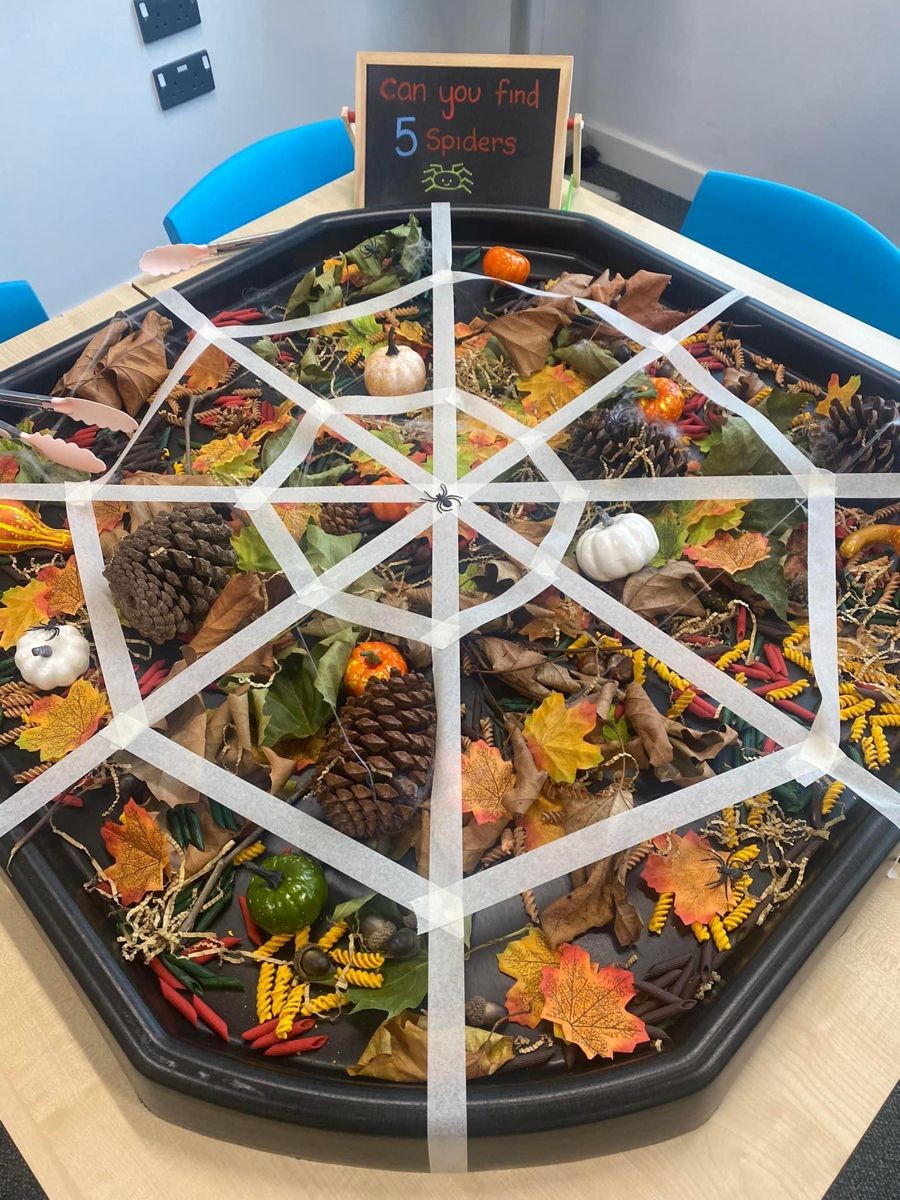 Een speeltafel met herfstbladeren, dennenappels, pompoentjes en vijf plastic spinnen op een spinnenweb van tape.