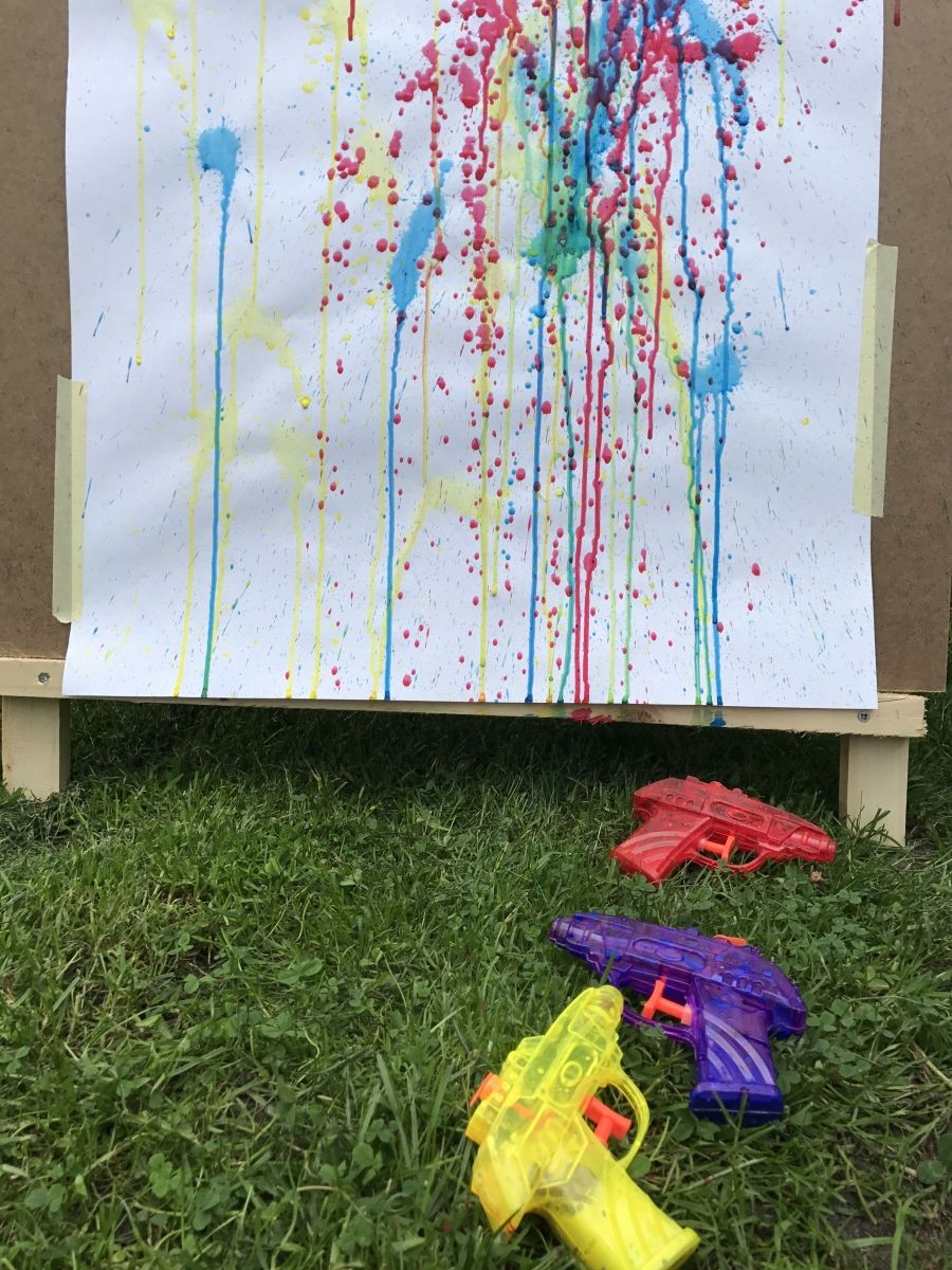 Een wit blad met verfspatten hangt op een bord, vooraan liggen drie kleurrijke waterpistolen in het gras.