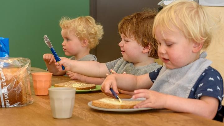 Drie jonge kinderen smeren boter op hun boterhammen aan een houten tafel.