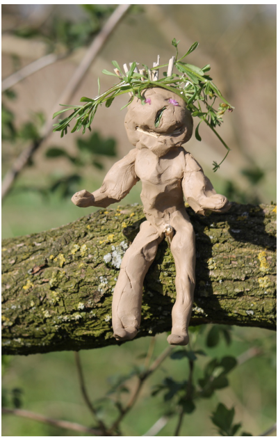 Een zelfgemaakte kleifiguur met planten op het hoofd zit op een boomtak in een buitenomgeving.