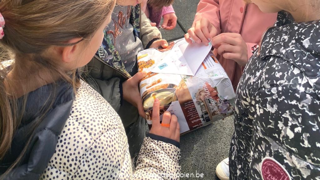 Vier kinderen staan dicht bij elkaar en openen een ingepakt cadeau buiten op het asfalt.