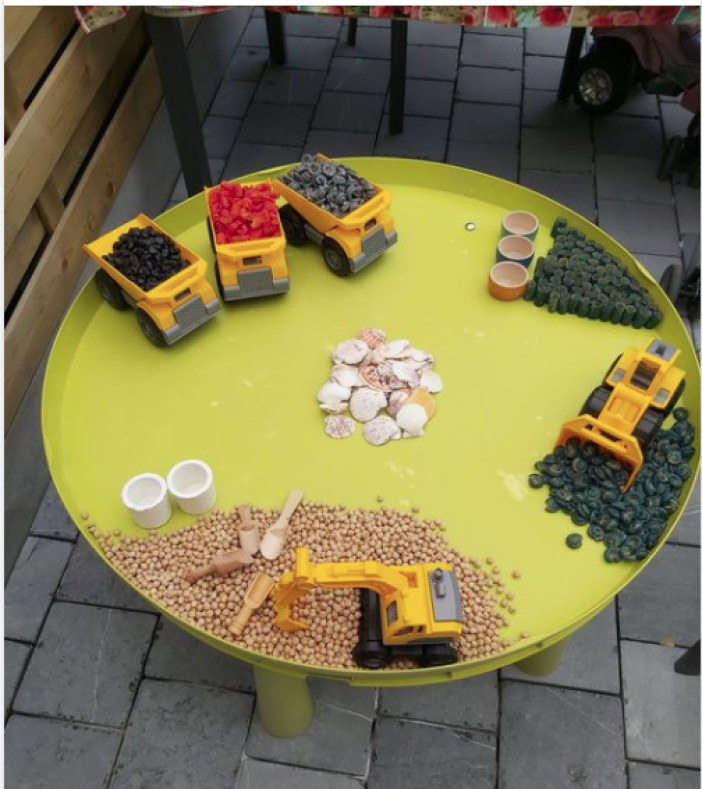Een groene tafel met speelgoeddumpers en -graafmachines vol met gekleurde knopen, houten kralen, schelpen en cups.