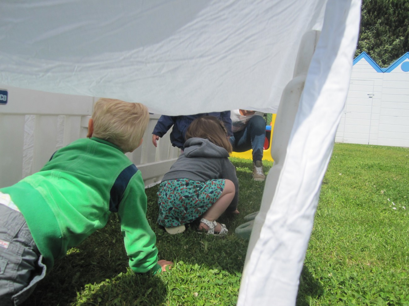 Drie kinderen kruipen onder een witte doek op het gras in een speeltuin.