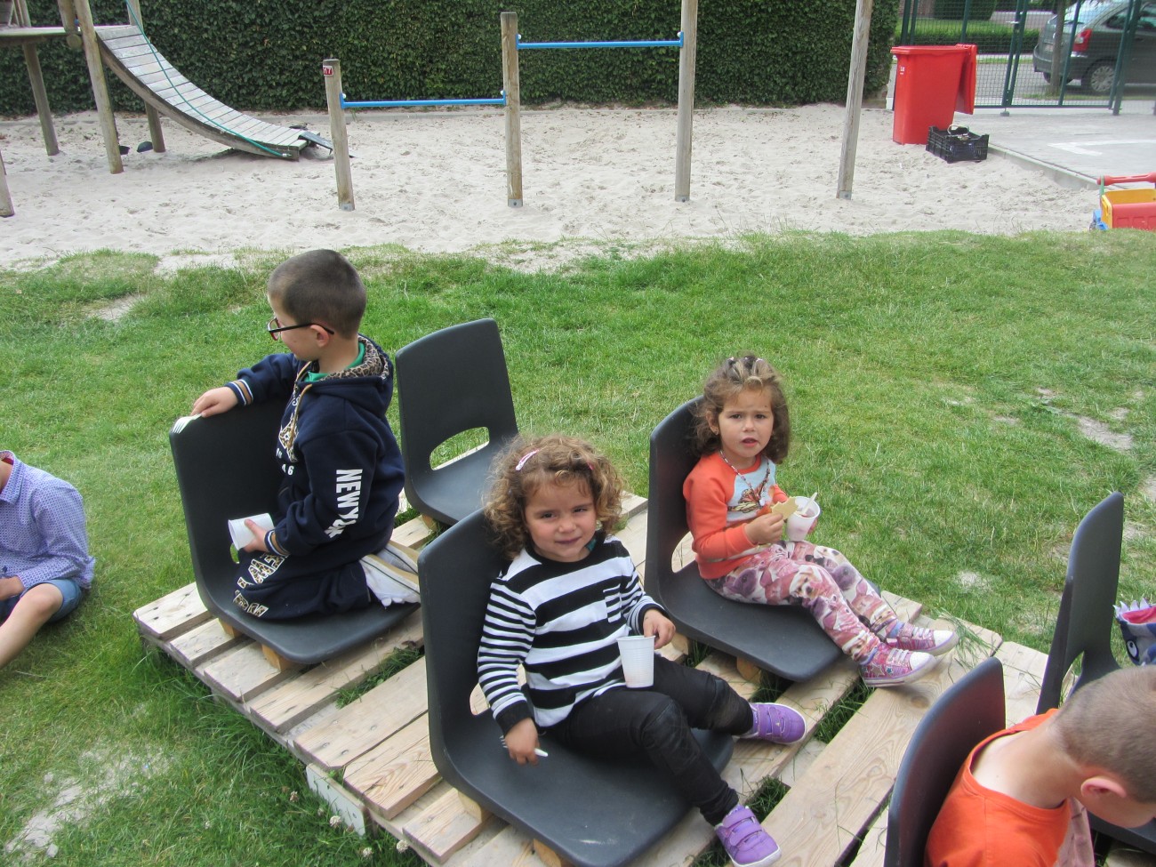 Drie kinderen zitten op stoelen op een houten platform in een grasrijke speelomgeving.