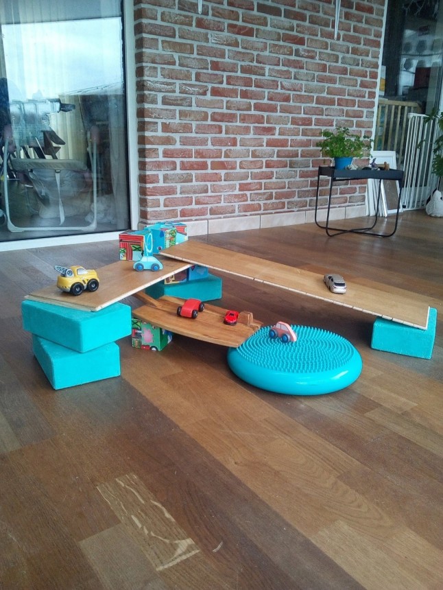 Een zelfgemaakte houten speeltuin met autootjes, plank en blauwe kussens staat op een houten vloer.