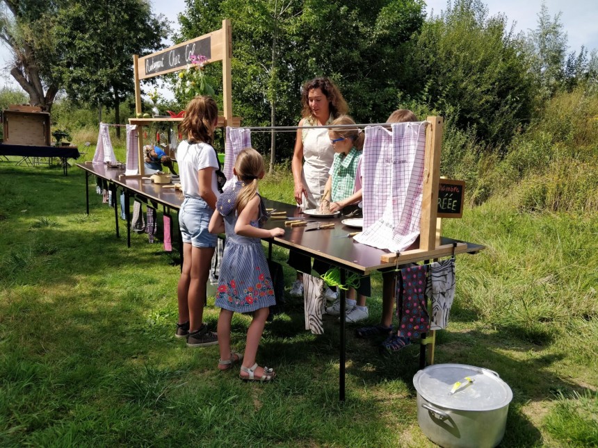 Kinderen staan buiten aan een lange tafel met doeken, onder begeleiding van een vrouw, bij een kraam in een groene omgeving.