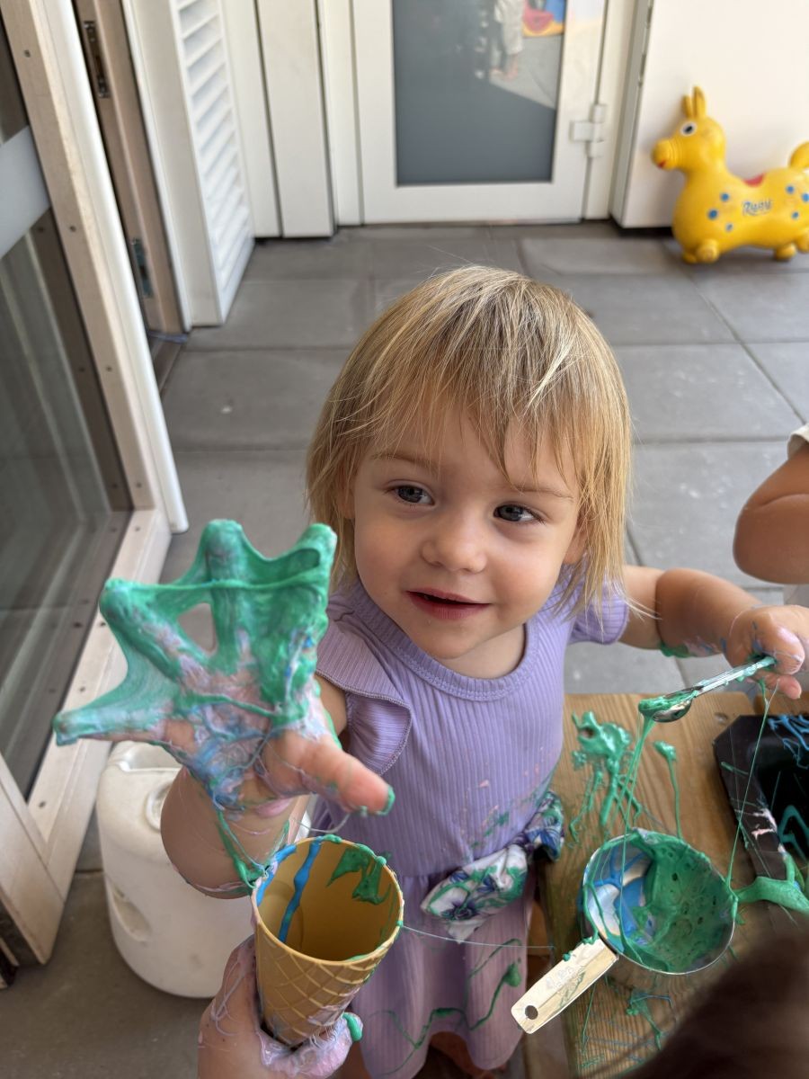 Een klein meisje speelt met groene slijm en houdt een ijsvorm en maatbekers vast, met een gele springkoe op de achtergrond.