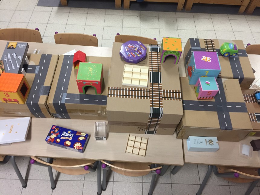 Een klaslokaaltafel met kartonnen dozen die met tape wegen, spoorrails en huizen voorstellen, en doosjes met snacks erop.