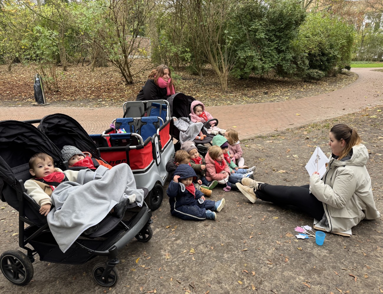 Een groep peuters zit op de grond terwijl een volwassen persoon hen voorleest in een park.