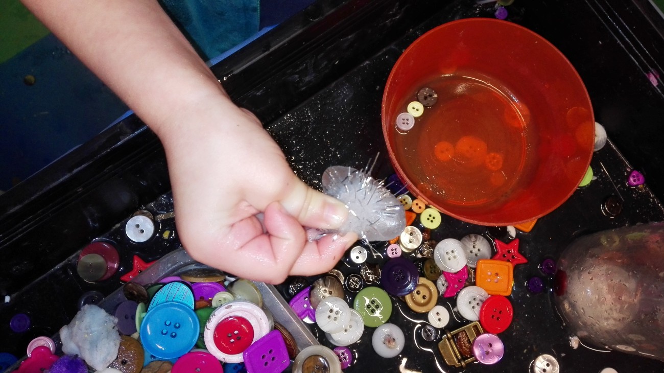 Een hand houdt een plastic zakje vast boven een bak met knopen en kleurrijke plastic items.