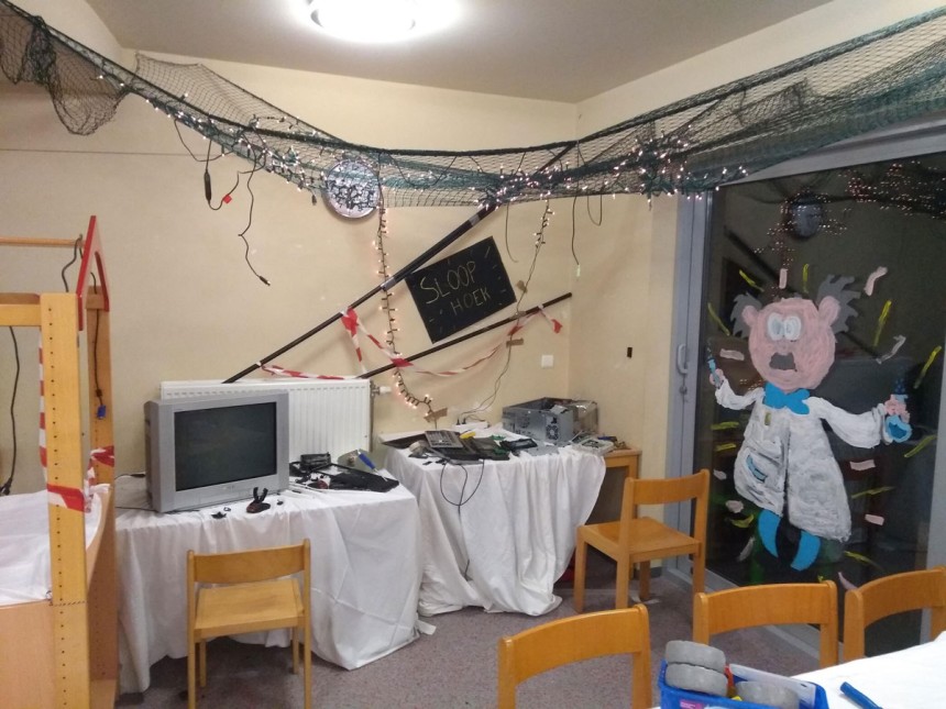 Een kamer met computerapparatuur op gedekte tafels, stoelen, een zwarte bord met tekst en een cartoon op het raam.