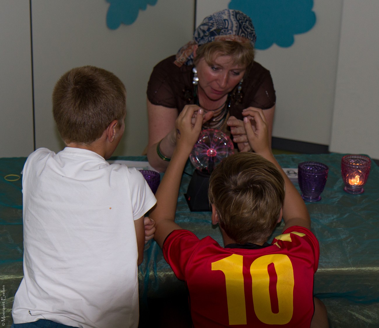 Twee kinderen kijken nieuwsgierig naar een vrouw die hen iets laat zien met een kristallen bol.