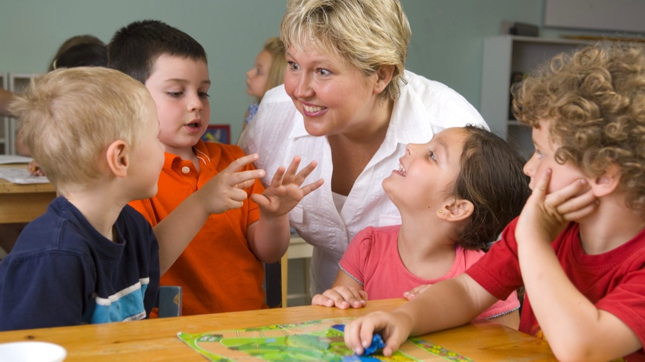 Een vrouw praat met vier kinderen aan een tafel met een bordspel in een klaslokaal.