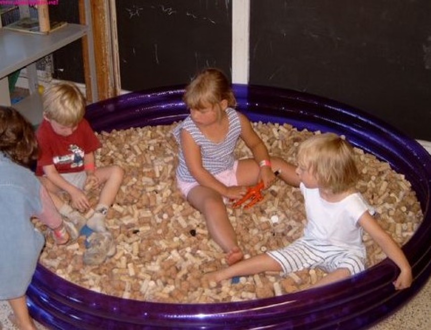 Drie kinderen zitten in een kinderzwembad gevuld met houten kurken, bezig met spelen.
