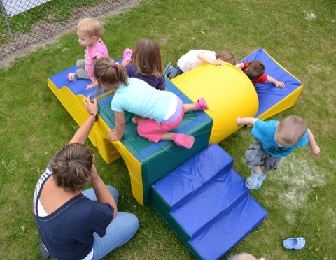 Eén volwassenen en meerdere kinderen spelen op een kleurrijk speeltoestel in een grasrijke omgeving.