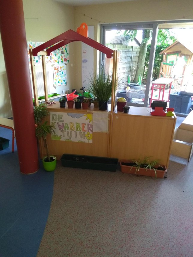 Speelhoek in een klaslokaal met planten, een houten kraam en uitzicht op een tuin buiten.