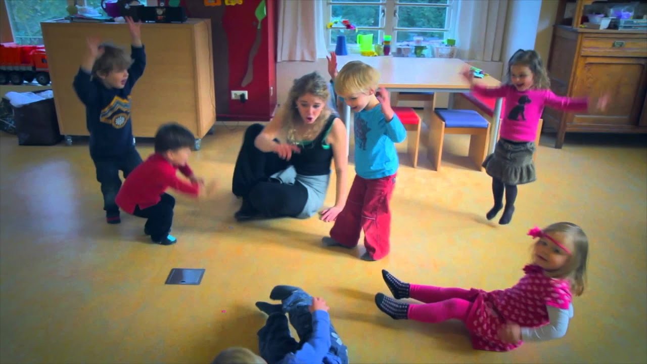 Een vrouw zit op de grond met vijf spelende kinderen in een kleurrijke kamer.