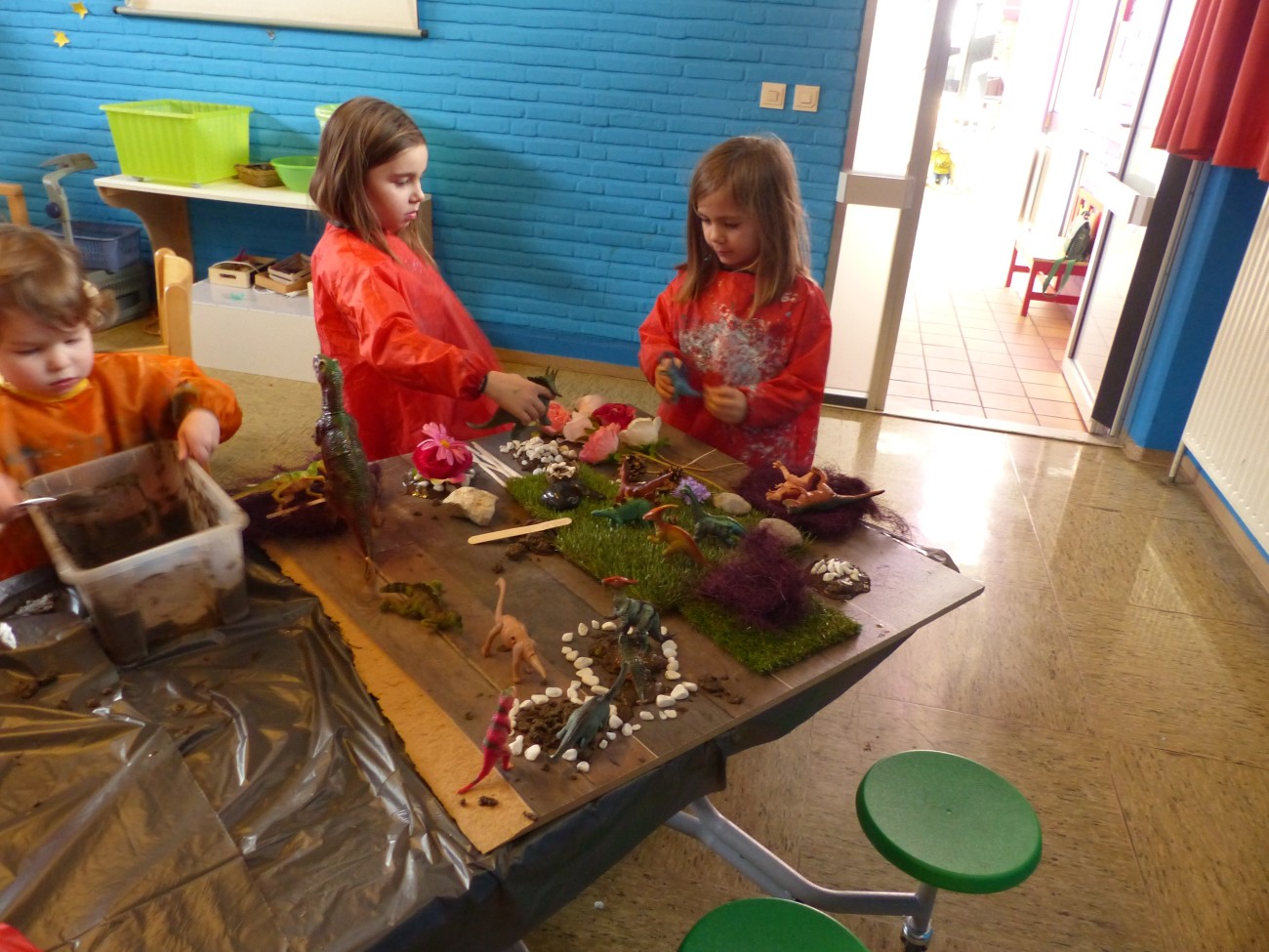 Drie kinderen in oranje schorten zijn bezig met knutselen aan een tafel met verschillende materialen.