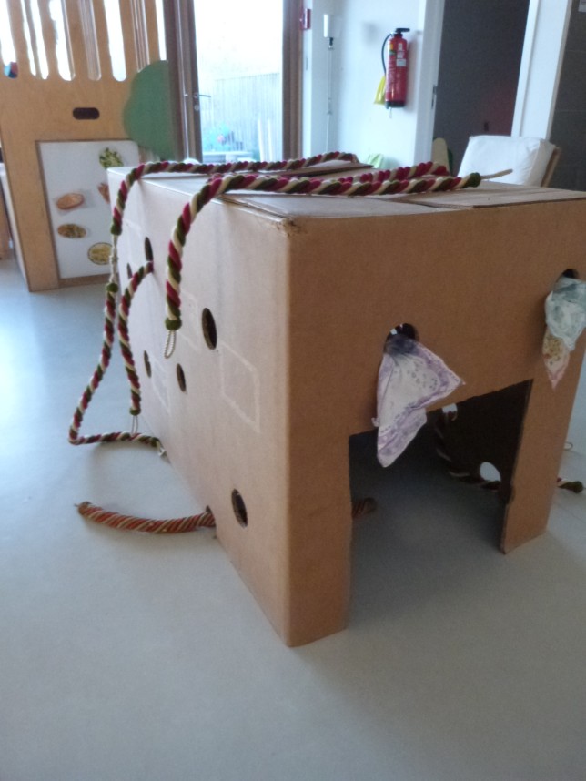 Een kartonnen dozenstructuur met openingen, touwen en een doekje op de vloer van een binnenruimte.