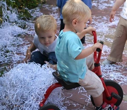 Twee kinderen spelen buiten, één fietst op een rode driewieler tussen stapels witte versnipperde papieren.