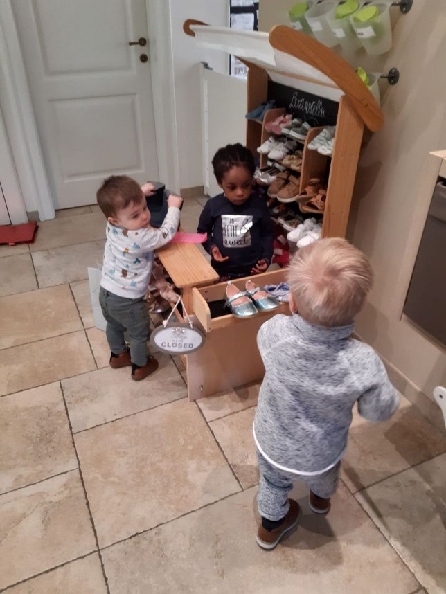 Drie jonge kinderen spelen bij een houten kraam met kinderschoenen in een gang.