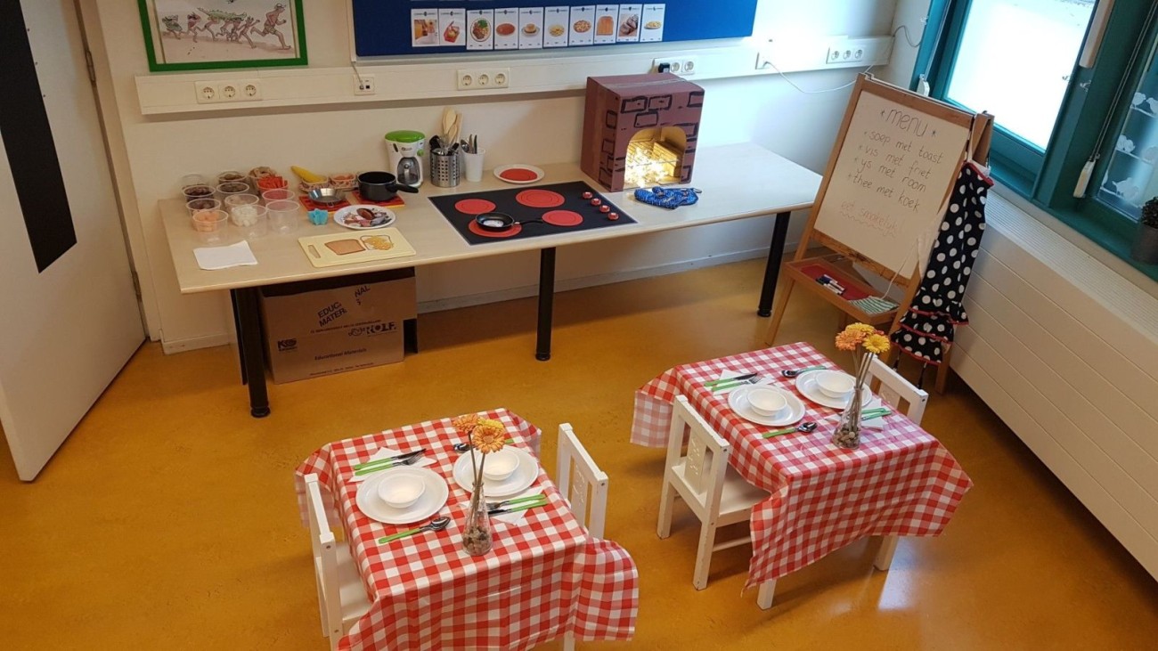 Speelhoek in een klaslokaal met kinderkeukentje, speeltafeltjes met rood-witte geruite tafelkleden en een menu op een schoolbord.