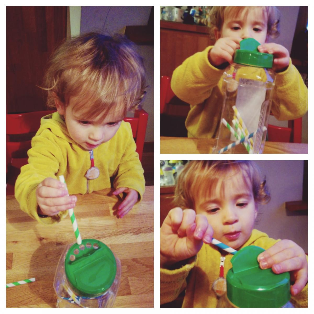 Een kind in een gele trui speelt met een glazen pot met een rietje aan een houten tafel.