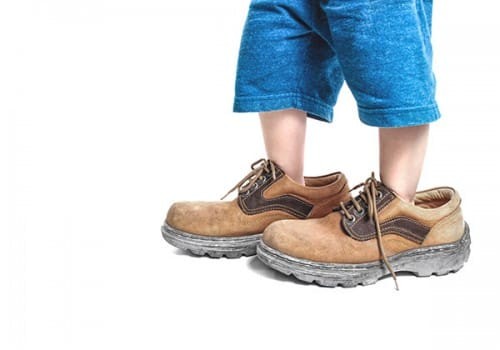 Kind met blauwe short draagt te grote bruine werkschoenen op witte achtergrond.