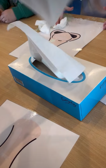Kinderen gebruiken een doos met tissues en hebben tekeningen met dierenkoppen op tafel liggen.