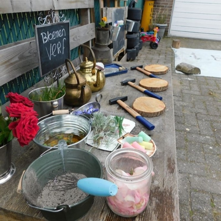 Tafel met bloemen, verf, houten schijven, hamers, theepotten en ander schildermateriaal buiten aan een muur.