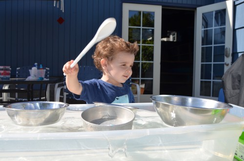 Een kind met bruin krullend haar speelt met een lepel in een waterbak met metalen kommen.
